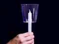 Hand holding a white candle fitted with a clear candle cup against a dark background.